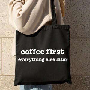 May include: Black tote bag with the white text "coffee first everything else later." The bag has a rectangular shape with two black handles. The bag is being held by a person.
