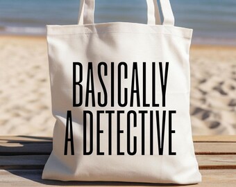 May include: A white tote bag with the text "BASICALLY A DETECTIVE" in black. The bag has two white handles and is sitting on a wooden surface. The background shows a beach with sand, ocean, and blue sky.