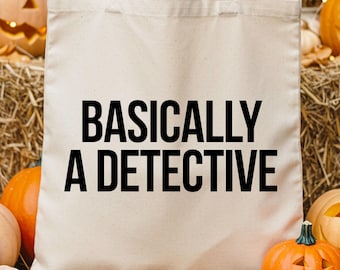 May include: A cream-colored tote bag with the text "BASICALLY A DETECTIVE" in black. The bag is held in front of a fall-themed display of pumpkins, hay bales, and autumn flowers. Some pumpkins are carved with jack-o'-lantern faces.