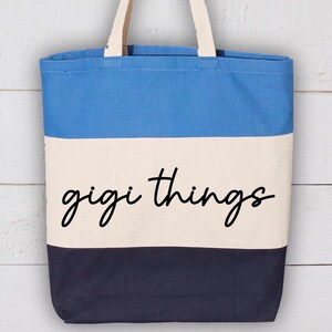 Op de afbeelding: Een canvas draagtas met een blauw, wit en marinekleurig blokontwerp. De woorden "gigi things" zijn in een zwart scriptschrift over de witte streep geschreven. De tas heeft crèmekleurige handvatten. Rechts staat een stengel met felroze bougainvillea bloemen.
