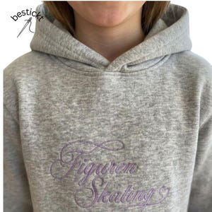 Könnte beinhalten: Ein graumelierter Kapuzenpullover mit dem gestickten violetten Schriftzug "Figuren Skating" und einem kleinen Herzsymbol. Der Hoodie hat eine dicke Kapuze und lange Ärmel. Das Logo "bestickt" mit Nadel und Faden ist oben links zu sehen.