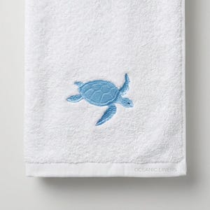 Könnte beinhalten: Weißes Handtuch mit hellblauem, gesticktem Meeresschildkröten-Design. Das Handtuch besteht aus einem weichen, saugfähigen Material und trägt unten den Text "OCEANIC LINERS".