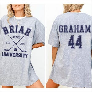 以下が含まれることがあります： グレーのBriar University Hawks Tシャツ。前面には「Briar University」の文字と、交差したホッケーのスティックとパックが描かれています。背面には「Graham」と数字の「44」がネイビーブルーで表示されています。
