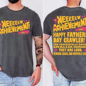 Puede incluir: Camiseta gris oscuro con el texto "NEEEEEW ACHIEVEMENT!" en amarillo y rosa. La parte trasera de la camiseta dice "HAPPY FATHERS DAY CRAWLER!" con texto adicional sobre la crianza de los hijos. Una camiseta divertida e informal.