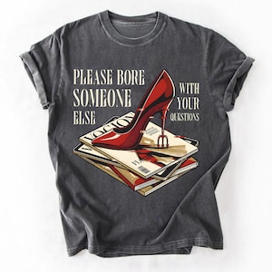 May include: Dark gray t-shirt featuring a red high-heeled shoe resting on a stack of magazines. The text on the shirt reads "PLEASE BORE SOMEONE ELSE WITH YOUR QUESTIONS."