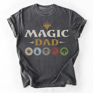 May include: Dark gray t-shirt with the text "MAGIC DAD" in white and gold. Below the text are six circular symbols in various colors. The shirt has rolled-up sleeves.