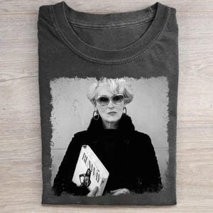 May include: Dark gray t-shirt featuring a black and white image of a woman wearing sunglasses and holding a magazine with the word "Runway" on it. The image has a distressed, vintage look.