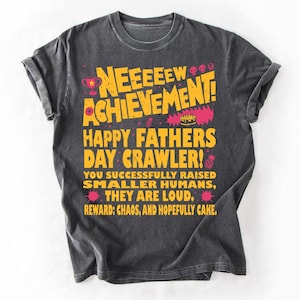 Puede incluir: Camiseta gris oscuro con el texto "NEEEEEW ACHIEVEMENT! HAPPY FATHERS DAY CRAWLER!" en amarillo y rosa. El mensaje de la camiseta celebra el Día del Padre y los desafíos de criar hijos, con un tono humorístico.