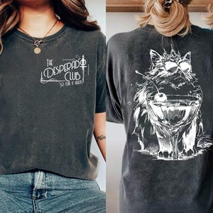May include: Dark grey t-shirts with white graphics. One shirt features "The Desperado Club" text. The other shirt has a cat wearing sunglasses and holding a martini glass with a cherry. Both shirts are short-sleeved.