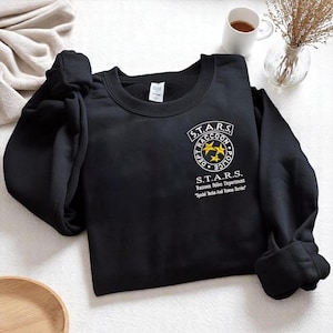 Könnte beinhalten: Schwarzes Sweatshirt mit dem S.T.A.R.S.-Logo der Raccoon Police Department. Das Logo ist weiß und goldfarben, mit dem Text "S.T.A.R.S." und "Raccoon Police Dept." Das Sweatshirt hat lange Ärmel und einen Rundhalsausschnitt.