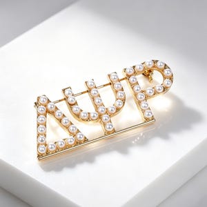 May include: A gold-toned metal brooch featuring the Greek letters Delta, Psi, and Phi, outlined with small white faux pearls. The letters are connected by a horizontal bar at the base, creating a structured, decorative accessory for clothing.