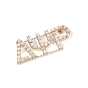 Peut inclure: Une broche en métal doré représentant les lettres grecques Delta, Psi et Phi, chacune ornée de petites perles blanches artificielles rondes. Les lettres sont reliées dans un design horizontal avec un mécanisme d'épingle visible au dos.