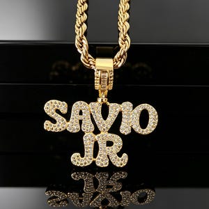 May include: A gold-toned rope chain necklace featuring a large, custom pendant that spells out the name SAVIO JR in capital letters. The pendant is encrusted with numerous small, clear, sparkling stones and hangs from a rectangular bail.