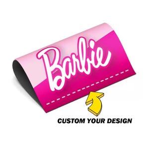 May include: A pink and white graphic with the word "Barbie" in a cursive font. The graphic has a gradient effect and a dashed line along the bottom. A yellow arrow points towards the text "CUSTOM YOUR DESIGN."
