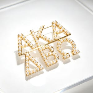 May include: A gold-toned metal brooch featuring the Greek letters Alpha, Kappa, Alpha, Delta, Beta, and Omega arranged in a stacked design. Each letter is outlined with small, round, white faux pearls embedded along the surface of the metal frame.