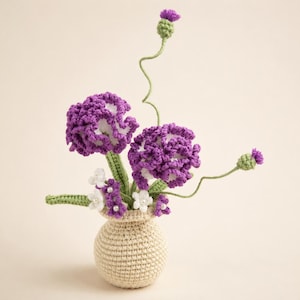 Puede incluir: Un arreglo de ganchillo con dos flores grandes con volantes en color morado y blanco, dos pequeños capullos morados tipo cardo en tallos verdes largos y flores blancas diminutas con centros de perlas, en un jarrón redondo de ganchillo beige.