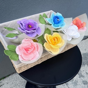May include: A collection of colorful, crocheted flowers arranged in a woven basket. The flowers include shades of purple, blue, pink, yellow, white, and orange, each with green leaves. The arrangement is presented on a black table.