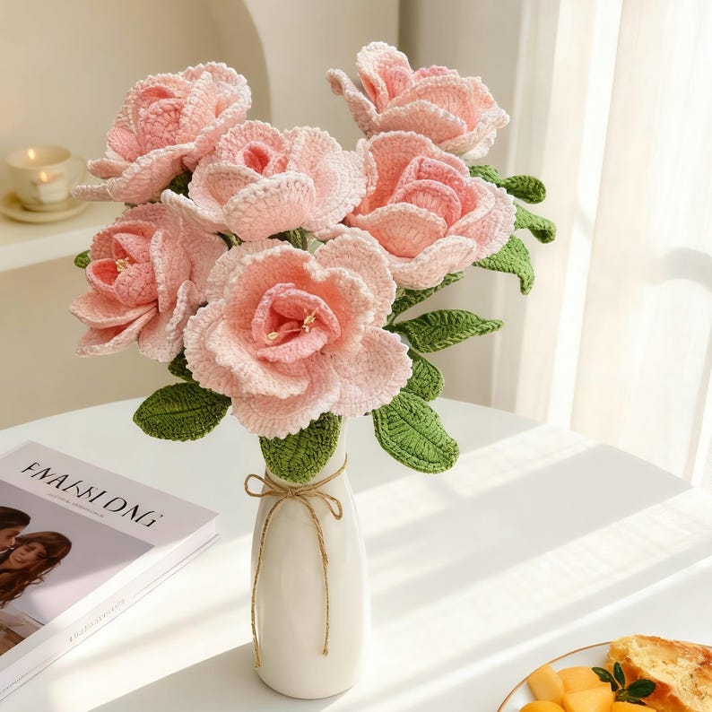 Puede incluir: Un ramo de flores de ganchillo hechas a mano en un jarr&oacute;n blanco. Las flores son de color rosa suave con hojas verdes, y el jarr&oacute;n est&aacute; atado con hilo. Las flores est&aacute;n dispuestas en un jarr&oacute;n sobre una mesa blanca. Se ve una revista.