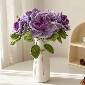Puede incluir: Un ramo de flores de ganchillo hechas a mano en un jarr&oacute;n blanco. Las flores son de varios tonos de morado, con hojas verdes. El jarr&oacute;n es alto y delgado, atado con un lazo de cuerda. Un art&iacute;culo de decoraci&oacute;n del hogar &uacute;nico.