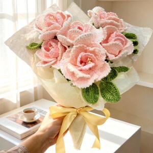 Puede incluir: Un ramo de flores de ganchillo rosas y verdes, envuelto en papel blanco y atado con una cinta amarilla. Un regalo &uacute;nico y hecho a mano, ideal para decoraci&oacute;n.