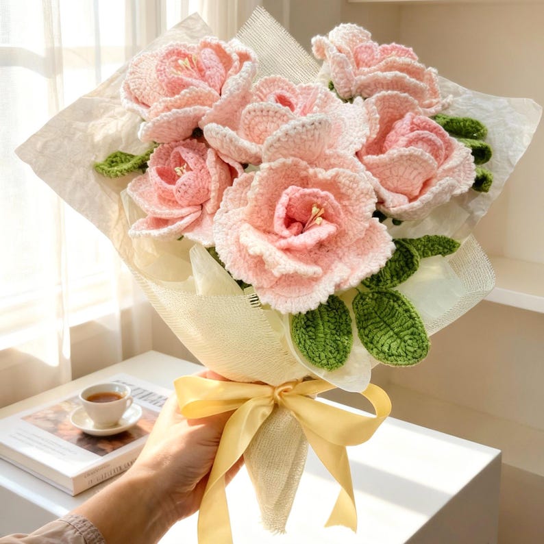 Puede incluir: Un ramo de flores de ganchillo rosas y verdes, envuelto en papel blanco y atado con una cinta amarilla. Un regalo &uacute;nico y hecho a mano, ideal para decoraci&oacute;n.