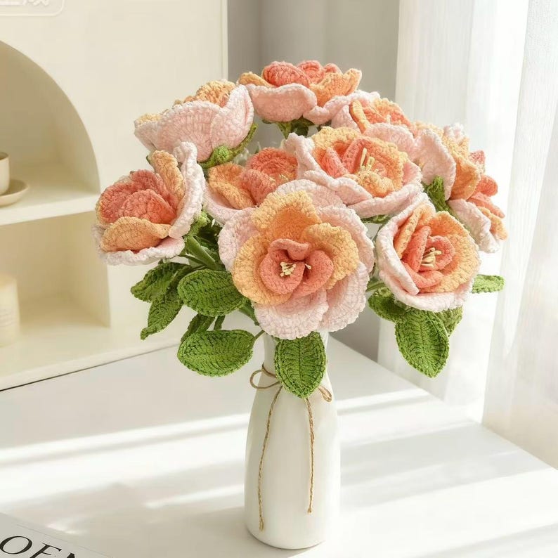 Puede incluir: Un ramo de flores de ganchillo en un jarr&oacute;n blanco. Las flores son en tonos melocot&oacute;n, naranja y rosa, con hojas verdes. El jarr&oacute;n est&aacute; atado con un lazo de cuerda. Las flores son un art&iacute;culo artesanal hecho a mano.