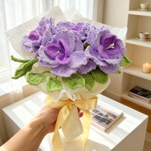 Puede incluir: Un ramo de flores moradas tejidas con hojas verdes, envuelto en papel blanco y atado con una cinta amarilla. Las flores hechas a mano tienen una textura detallada y est&aacute;n dispuestas para parecerse a las hortensias.