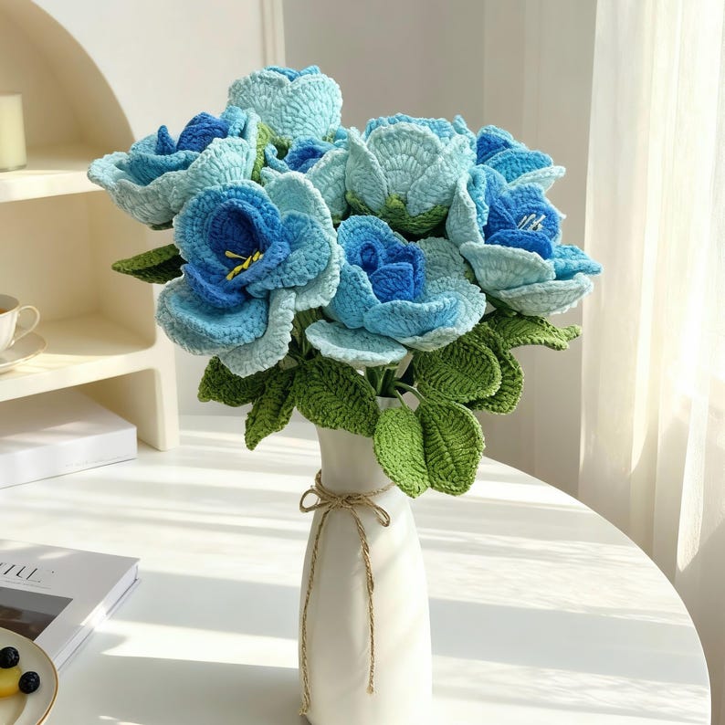 Puede incluir: Un ramo de rosas azules hechas a mano con ganchillo, con hojas verdes, presentado en un jarr&oacute;n blanco atado con hilo. Las rosas presentan diferentes tonos de azul. Un art&iacute;culo de decoraci&oacute;n &uacute;nico y hecho a mano.