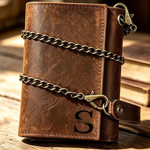 Puede incluir: Una cartera de cuero marrón de tres pliegues con textura desgastada, una cadena de metal color latón sujeta con un mosquetón y una lengüeta de cuero con botón a presión. El frente tiene grabada una letra S grande y el nombre Scott debajo.