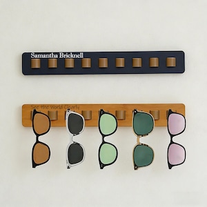 Puede incluir: Dos organizadores de gafas de sol, uno azul marino y otro de bambú, montados en una pared blanca. El organizador azul marino tiene el nombre "Samantha Bricknell" impreso. El organizador de bambú tiene el texto "See the World Clearly". Se exhiben cinco pares de gafas de sol.