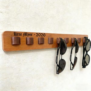 Puede incluir: Un soporte de madera para gafas de sol con lazos de cuero marrón y tres pares de gafas de sol colgando. El soporte tiene grabado el texto "Kree Myer • 2026". El soporte está montado en una pared blanca.