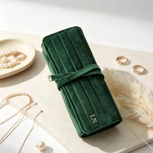 May include: A dark green velvet jewelry roll featuring vertical quilted stitching, a wrap-around tie closure with a metallic accent, and the embroidered initials LN on the front flap, designed for organizing and storing various jewelry items.