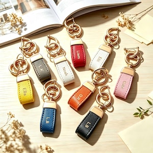 May include: A collection of nine rectangular leather keychains in various colors including yellow, gray, white, red, orange, blue, black, and pink. Each keychain features a gold-toned metal clasp and is personalized with a name or initials.