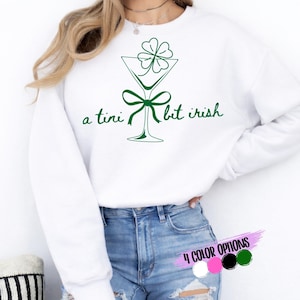 May include: White sweatshirt with a green martini glass design, a shamrock, and the text "a tini bit irish". The design includes a bow and the words are in a script font. The image also shows color options: white, pink, black, and green.