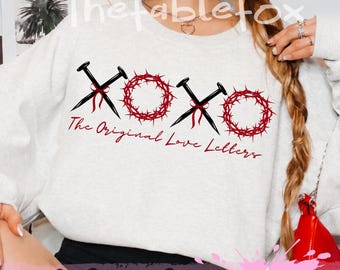 May include: A light gray crewneck sweatshirt featuring a graphic design with the text XOXO and The Original Love Letters. The design includes two black crossed nails tied with red ribbons and two red circular crowns of thorns.