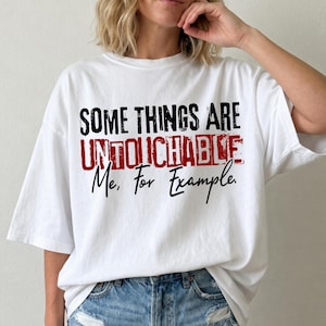 May include: White t-shirt featuring the quote "SOME THINGS ARE UNTOUCHABLE Me, For Example." The text is in black and red, with a distressed effect on "UNTOUCHABLE." The shirt has a casual fit and short sleeves.