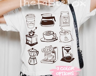 May include: A white t-shirt featuring a brown graphic print of nine coffee-related items, including a cup, a carafe, a bag labeled Fresh Roast, a grinder, a mug, a coffee bean sack, a moka pot, an espresso machine, and a pour-over.