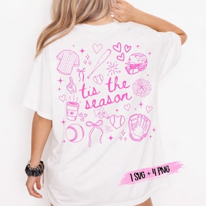 May include: White t-shirt with a pink baseball-themed graphic on the back. The design includes a baseball jersey, bat, helmet, glove, ball, and the phrase "tis the season". Additional elements include hearts, bows, and a coffee cup.