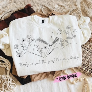 May include: White sweatshirt with a black line drawing of an open book, flowers, and a butterfly. The text "There's no such thing as too many books" is below the drawing. The bottom right corner has a pink banner that says "4 COLOR OPTIONS".