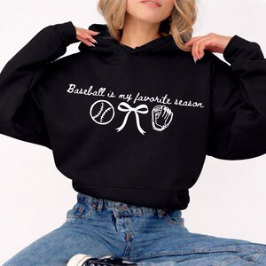 May include: Black hoodie with white text and baseball-themed graphics. The text reads "Baseball is my favorite season" with a baseball, bow, and baseball glove graphic. A casual, comfortable sweatshirt.