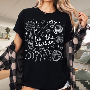 May include: Black t-shirt with white baseball-themed graphics and the text "tis the season." The design includes a baseball jersey, bat, helmet, glove, ball, and hearts. The shirt is a casual, everyday wear item.