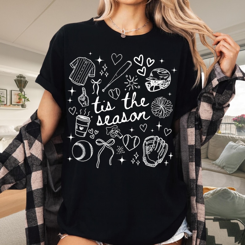 May include: Black t-shirt with white baseball-themed graphics and the text "tis the season." The design includes a baseball jersey, bat, helmet, glove, ball, and hearts. The shirt is a casual, everyday wear item.