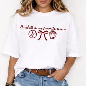 May include: White t-shirt with the text "Baseball is my favorite season" in a burgundy script. The design includes a baseball, a bow, and a baseball glove in burgundy. The shirt is a casual, everyday wear item.