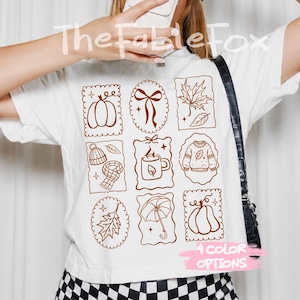 May include: A white t-shirt featuring a brown line-art grid of nine autumn-themed icons including a pumpkin, a bow, a maple leaf, a beanie, a scarf, a mug, a sweater, an oak leaf, and an umbrella. The text TheFableFox and 4 COLOR OPTIONS appear.
