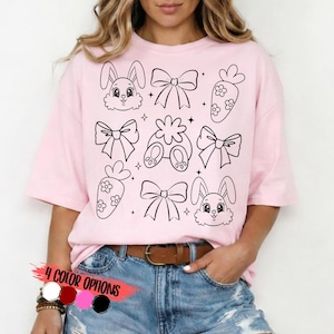 May include: A light pink t-shirt featuring a black and white Easter-themed design. The design includes bunny faces, bows, carrots, and bunny feet. The text "4 COLOR OPTIONS" is displayed at the bottom of the shirt, with color swatches.