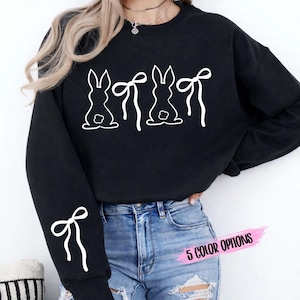 May include: Black sweatshirt with a white graphic of three bunny silhouettes with bows. A white bow is also on the left sleeve. The text "5 COLOR OPTIONS" is in pink. This sweatshirt is perfect for Easter.