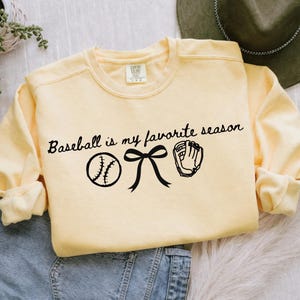 May include: A light yellow sweatshirt with the text "Baseball is my favorite season" in black script. The design includes a baseball, a bow, and a baseball glove. The sweatshirt is a crew neck style.