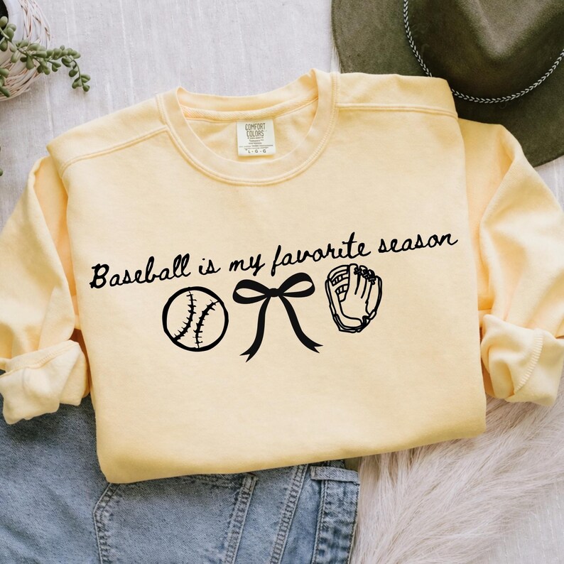 May include: A light yellow sweatshirt with the text "Baseball is my favorite season" in black script. The design includes a baseball, a bow, and a baseball glove. The sweatshirt is a crew neck style.