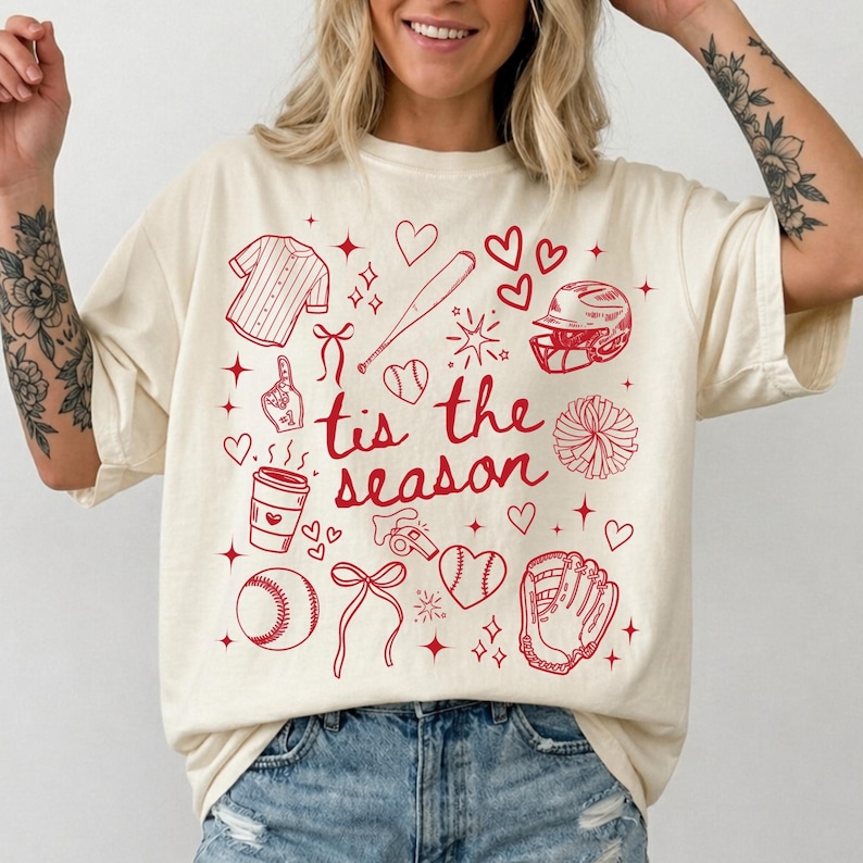 May include: Cream-colored t-shirt with a red baseball-themed graphic. The design includes a baseball jersey, bat, helmet, baseball, glove, and the phrase "tis the season". Hearts and stars are scattered throughout the design.