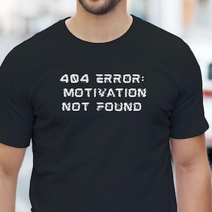 May include: Black t-shirt with the text "404 ERROR: MOTIVATION NOT FOUND" in white. The shirt is a crew neck style and is being worn by a person. The background is a blurred outdoor scene.